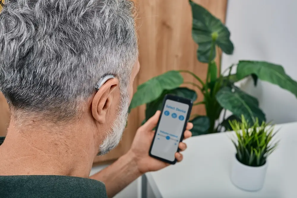 An older man with hearing impairment adjusts the settings on his behind-the-ear hearing aid.