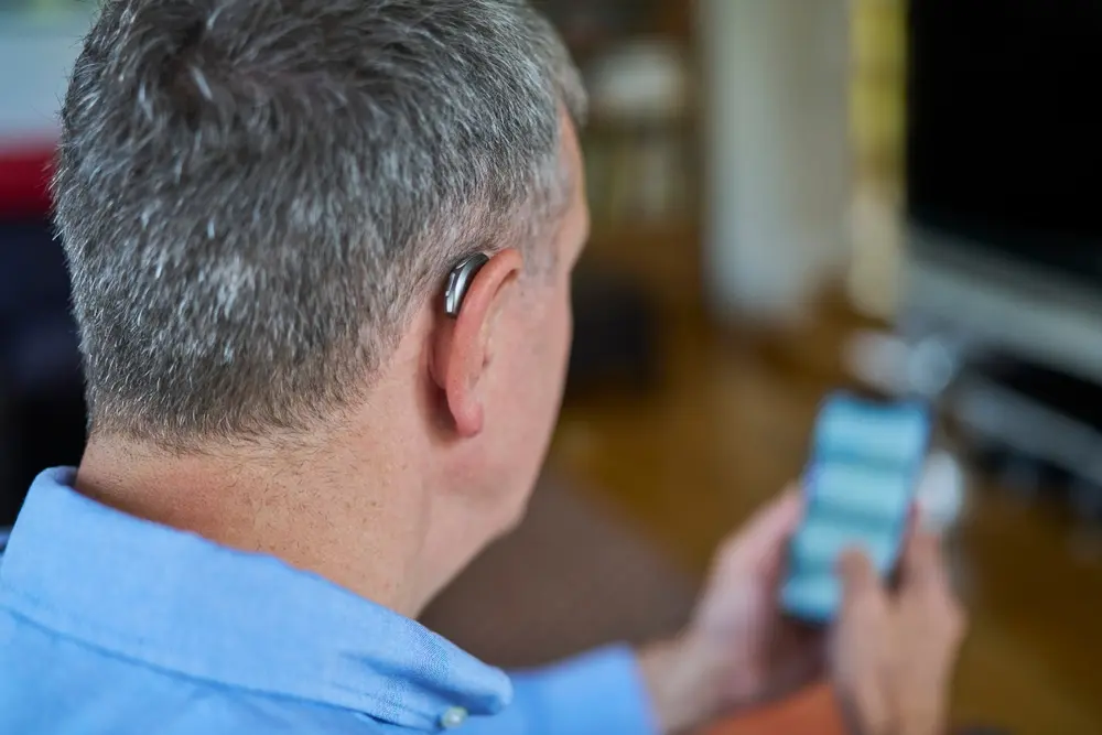 Man Wearing Wireless Hearing Aid Using Mobile Phone At Home.